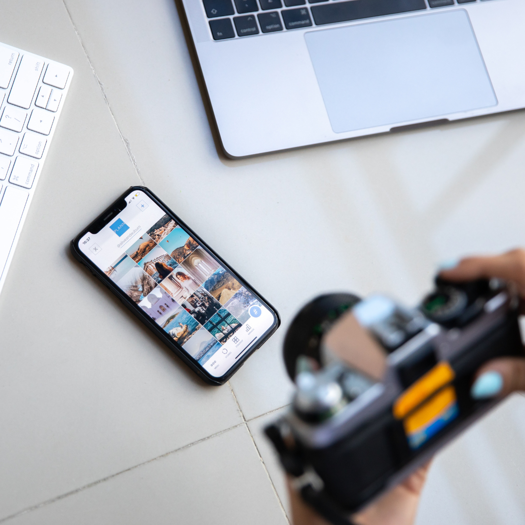 Social Media Content Erstellung: Fotograf hält eine Kamera, während auf einem Smartphone Instagram Fotos angezeigt werden. Im Hintergrund liegt ein Laptop und eine Tastatur auf einem modernen Schreibtisch.